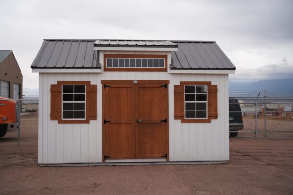 A 12x16 Studio Gable metal roofed storage shed with wooden doors and window trim, surrounded by a cloud-filled sky and several vehicles in the background.