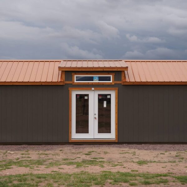 Front view of a large Studio Gable 12x28 storage shed with residential-style double doors, grey wood siding, and a metal roof.