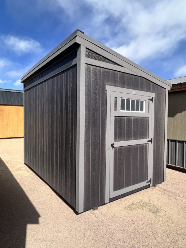 The sky is dotted with clouds as a 8x12 Urban Style storage shed with an angled roof stands tall, displaying the side marked by a grand door.