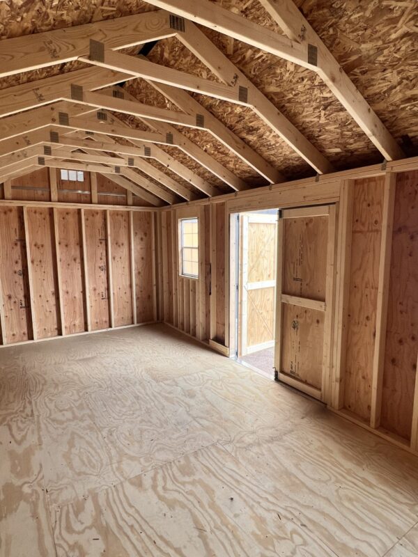 A beam of light illuminates the hardwood planks and lumber of the ceiling, walls, and floor of the 10x20 Gable Style storage shed.