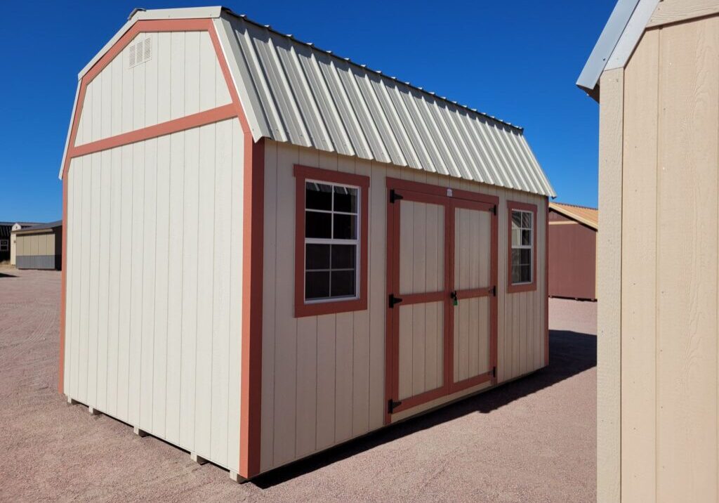 Barn style storage shed with double doors and two windows. Tan metal roof and walls with red trim.